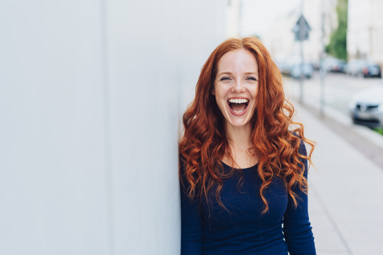 Red-haired woman with dental implants smiles while standing against a white wall