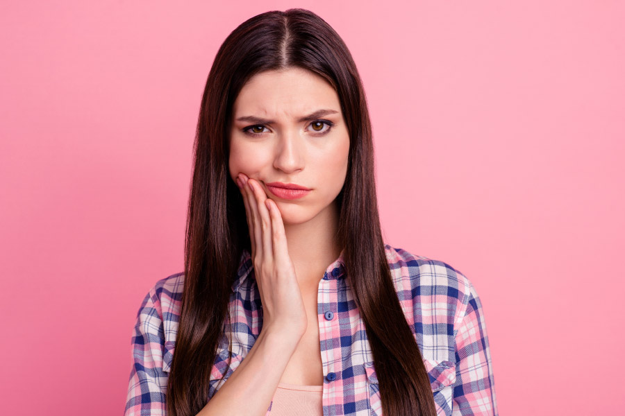 Brunette woman in a plaid shirt cringes in pain and touches her cheek because she needs an emergency dentist